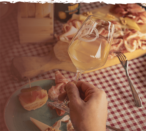 Mano che regge un calice di vino bianco naturale, con tagliere di salumi su tavola a quadretti.